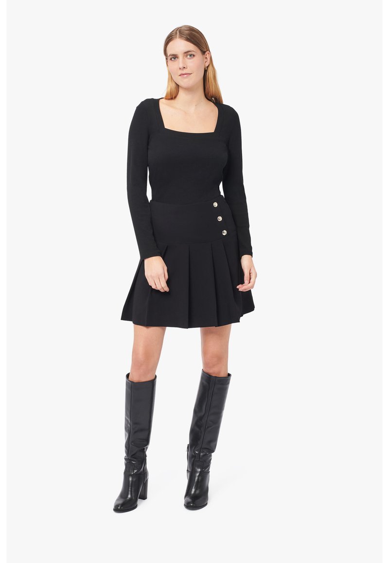 Schwarzes Langarmoberteil mit quadratischem Ausschnitt, kombiniert mit einem schwarz gefältelten Rock, der an der Seite drei silberne Knöpfe hat. Schwarze kniehohe Stiefel.