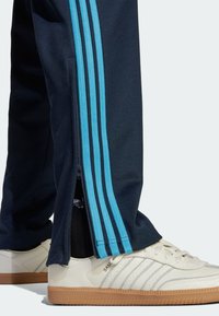 Navy träningsbyxor med turkosa sidostrecker, med en dragkedja vid fållen, i kombination med vita sneakers och brun gummisula.