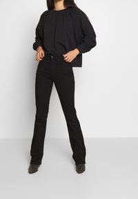 Blouse noire à manches longues avec un col rond et un design plissé, accompagnée de jeans évasés noirs taille haute et de chaussures sombres.