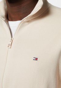 Primo piano di una persona che indossa una giacca beige con cerniera sopra una maglietta bianca, con un piccolo logo rosso, bianco e blu navy sul petto.