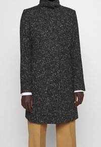 Svart- och vitmönstrad jacka med hög krage, långa ärmar och rak skärning, matchad med beiga byxor. Slitstark tyg och skräddarsydd passform.