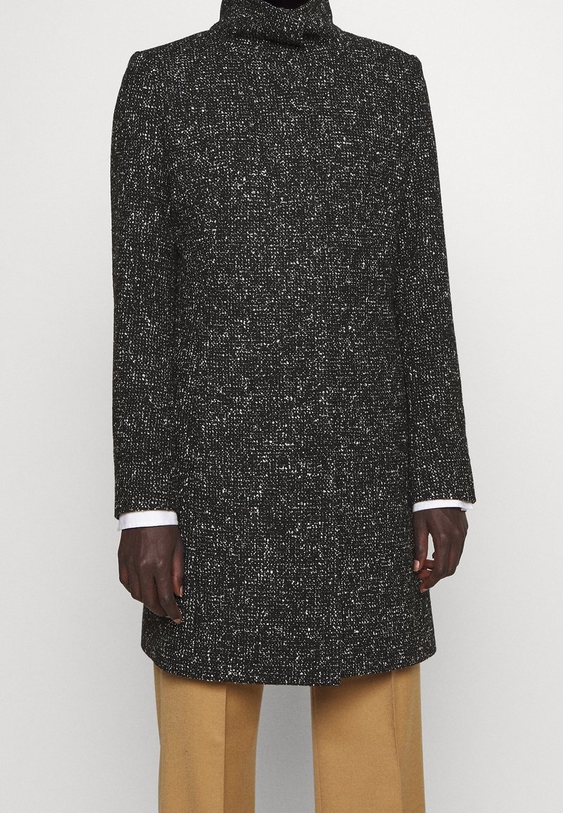 Svart- och vitmönstrad jacka med hög krage, långa ärmar och rak skärning, matchad med beiga byxor. Slitstark tyg och skräddarsydd passform.