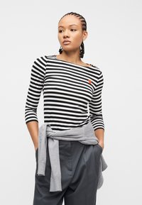 Νεαρή γυναίκα με πλεξούδες, που φοράει μπλούζα με ασπρόμαυρες ρίγες και μικρή κόκκινη καρδιά, γκρι πουλόβερ δεμένο στη μέση και σκούρο παντελόνι.