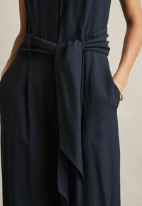 Marineblauwe jumpsuit met een gestrikt taille-detail, voorzien van zakken en een ontspannen pasvorm. Glad fabric met een lichte glans en subtiele textuur.