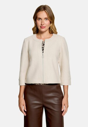 Femme portant une veste crème zippée avec manches trois quarts par-dessus un haut imprimé léopard et un pantalon en cuir marron foncé.