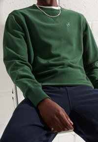 Osoba v zelené mikině s malým logem a stříbrným řetízkem, sedící s rukama položenýma na tmavě modrých kalhotách.