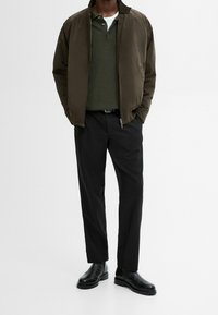 Bruine geïsoleerde jas, olijfgroene polo, zwarte broek en zwarte laarzen. Gladde stof, gestructureerde pasvorm en minimale ritsaccenten.