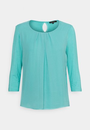 Licht turquoise blouse met driekwart mouwen, plooien onder de ronde halslijn en een kleine sleutelgatuitsteker bij de achterkraag.