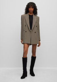 Blazer doppiopetto in un motivo houndstooth marrone e nero, con spalle strutturate. Abbinato a un top aderente e stivali in suede neri al ginocchio.