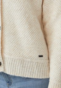 Cremefarbener Strickpullover mit strukturiertem Rautenmuster, geripptem Saum und kleinem schwarzen Metalletikett, getragen zu blauen Jeans.