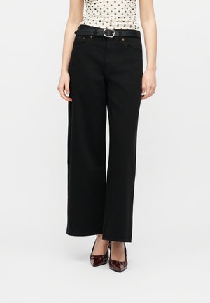 Femme portant un pantalon noir taille haute à jambes larges avec une ceinture noire, un haut sans manches à pois, et des talons pointus marron foncé, debout devant un fond blanc.