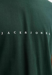 Σκούρο πράσινο ύφασμα με λευκό κείμενο "JACK & JONES" εκτυπωμένο οριζόντια στη μέση.