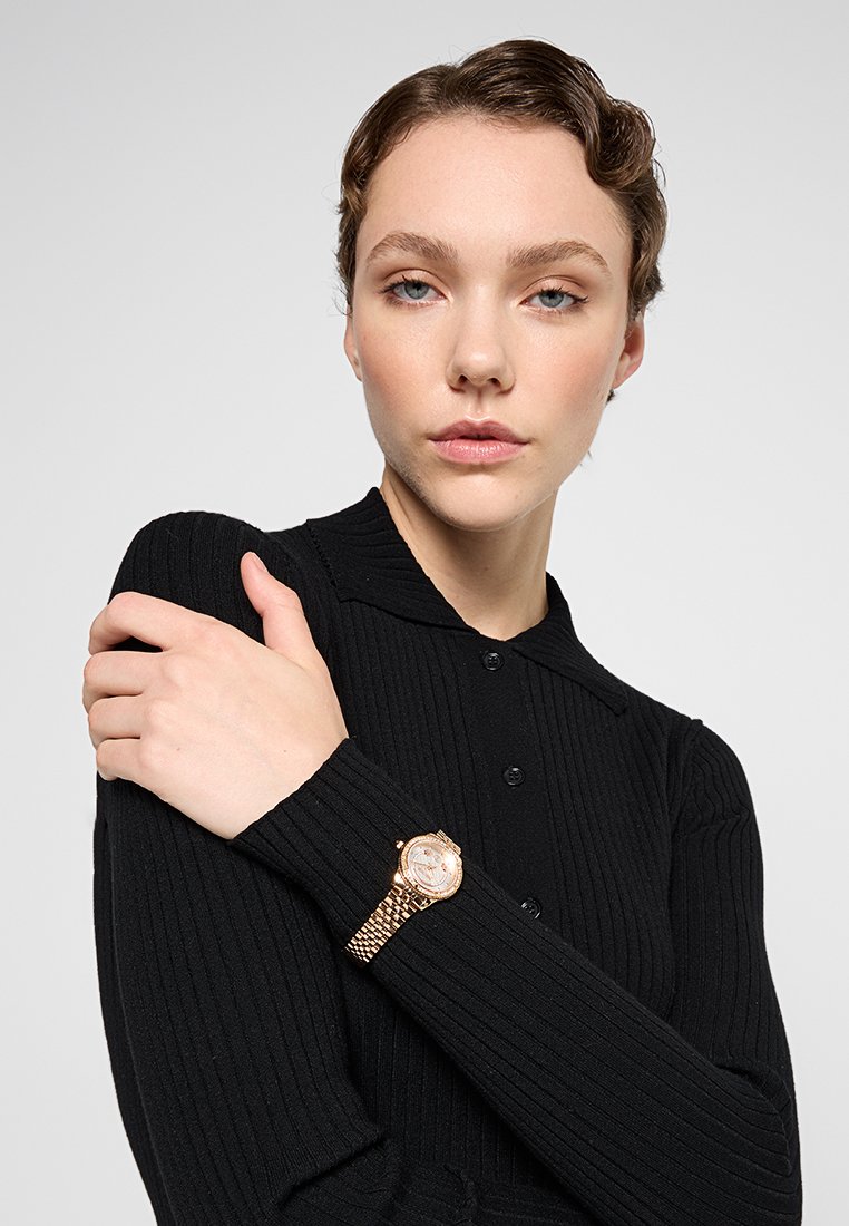 Montre en or avec un bracelet texturé, cadran rond et marqueurs numériques, portée au poignet d'un modèle. Haut à col côtelé noir.