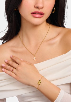 Frau mit goldener Kreuzkette, goldenem Rosenarmband, Rubinring und goldenen Creolen, mit zart rosa Nagellack auf nackten Schultern.