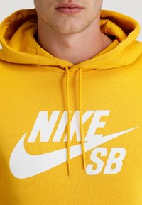 Närbild av en person som bär en gul hoodie med vit text "NIKE SB" och swoosh-logga på bröstet, med dragsko i huvan.