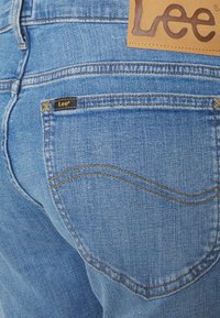 Jeans de ganga azul com um acabamento texturizado, apresentando uma etiqueta de pele castanha e uma pequena etiqueta preta com "Lee" no bolso de trás.