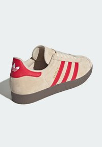 Beige mockaskors med tre röda ränder, guld "Gazelle"-logotyp och en röd hälflik med vit logotyp. Gummisula, rund tånäs.