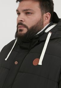 Sort polstret jakke med hætte, der har hvide snore, et læderlogo-patch og en quiltet tekstur.