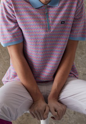 Pikepaita lyhythihaisena, jossa on vaaleanpunainen ja sininen sahalaita-kuvio, varustettuna vetoketjukauluksella ja pienellä mustalla logolla rinnassa.