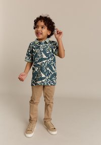 Korte mouwen polo shirt met een groen tropisch bladpatroon, marineblauwe basis en gestreepte kraag. Gekleed met beige broek en lichte sneakers.