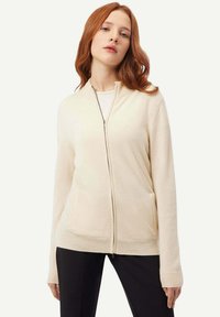 Krämfärgad, zip-up cardigan i mjukt stickat tyg med långa ärmar, två framfickor, ribbade ärmslut och nederkant. Maskintvättbar för lätt skötsel.