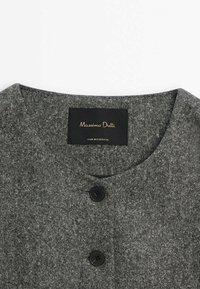 Harmaan tekstuurilla varustettu kangas ylhäältä, jossa on pyöreä pääntie, kahdella mustalla napilla ja etiketissä lukee "Massimo Dutti, valmistettu Portugalissa."