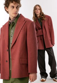 Dwa modele prezentują oversize'ową, czerwono-brązową wełnianą marynarkę z klapami, przednimi kieszeniami i guzikami, w zestawieniu z kontrastującymi strojami.