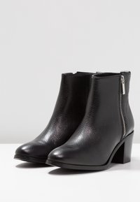 Stivaletti neri in pelle con finitura testurizzata, punta rotonda, zip laterale e tacco alto chunky, posizionati su uno sfondo neutro.