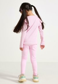 Lyserød langærmet skjorte med flæsedetaljer og matchende tætsiddende bukser. Lyse sneakers med grønne accenter. Mørkt, glat hår sat op i to hestehaler.