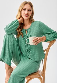 Completo da casa verde chiaro con trama, composto da top con nodo frontale e pantaloni larghi. Modella tiene in mano una tazza bianca, seduta su una sedia di legno.