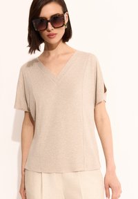 Beżowa koszula z dekoltem w serek i krótkim rękawem, wykonana z miękkiego materiału. Charakteryzuje się luźnym krojem i dyskretnymi szwami, co zapewnia schludny wygląd.
