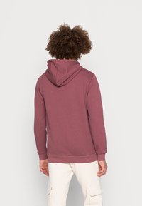 Felpa con cappuccio color mauve, dotata di cappuccio con cordino, polsini a costine e una finitura liscia e strutturata. Indossata con pantaloni chiari.