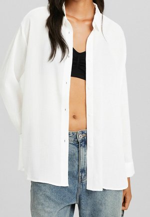 Femme portant une chemise blanche à manches longues ouverte sur un crop top noir et un jean bleu délavé, debout devant un fond gris clair.