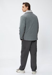 Veste matelassée grise avec un col en polaire gris, portée sur un pantalon cargo noir et des baskets crème, vue de dos sur un fond uni.