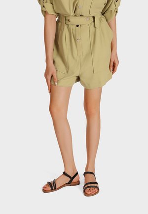 Donna che indossa pantaloncini beige con cintura e sandali neri bassi con cinturini, in piedi davanti a uno sfondo bianco uniforme.
