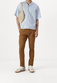 Jasnoniebieska koszula w paski z krótkim rękawem, beżowe spodnie, kremowe trampki z czarnymi akcentami oraz beżowa torba na ramię.