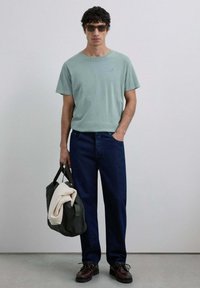 Lichtgroene katoenen T-shirt, donkerblauwe denimjeans, bruine veterschoenen, houdt een donkergroene tas vast met een crèmekleurige trui erin.