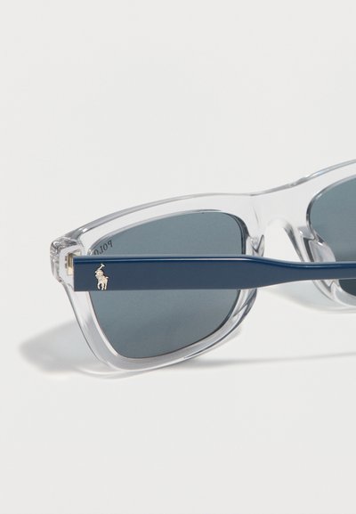 Lunettes de soleil avec montures transparentes, verres foncés et branches bleu marine ornées d'un petit emblème doré d'un joueur de polo.