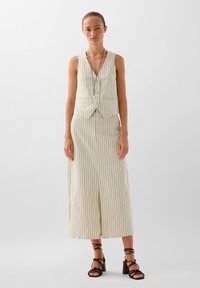 Beige gestreepte mouwloze vest met een V-hals en zilveren knopen, gecombineerd met een bijpassende lange rok met een split aan de voorkant. Strappy zwarte hakken.
