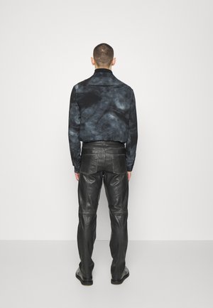 Mężczyzna w czarno-szarej kurtce typu tie-dye i czarnych skórzanych spodniach stoi przodem do gładkiej białej ściany.
