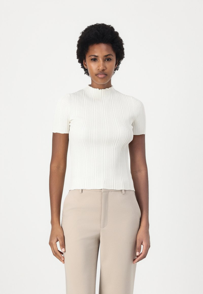 Light ribbed knit top in off-white with a high, ruffled neckline and short sleeves, paired with beige tailored trousers.