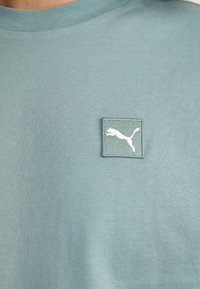 Tricou din bumbac verde deschis cu guler rotund, decorat cu un mic patch brodat alb reprezentând o felină săltând. Textură netedă.