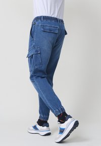 Modre denim jogger hlače s elastičnim pasom, cargo žepi in poševnimi robovi, v kombinaciji z belimi in modrimi športnimi čevlji.