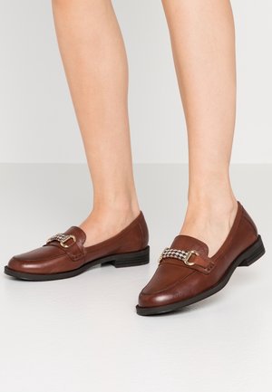 Brune læder loafers med guldfarvet hardware og hundetandsstofdetalje, båret på bare fødder, stående på en lysefaret gulv.