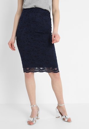 Femme portant une jupe crayon en dentelle bleu marine avec un motif floral et un ourlet festonné, associée à des sandales à talons hauts argentées et un haut gris.