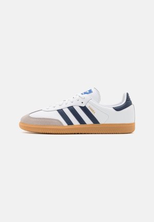 Biały sneaker Adidas Samba z granatowymi paskami, gumową podeszwą, beżowym noskiem i złotym napisem "SAMBA" z boku.