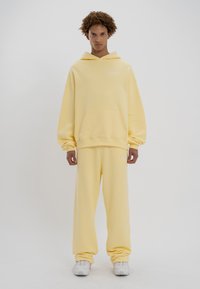 Sudadera amarilla con capucha y pantalones de chándal hechos de una tela suave, con un amplio bolsillo frontal y puños acanalados. Detalle de logo mínimo en el pecho.