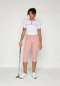 Ženska stoji z golfsko palico, oblečena v belo in roza polo majico, roza capri hlače in bele športne čevlje pred enotno ozadje.