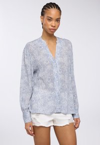 Camicia blu con motivo paisley, maniche lunghe, scollatura a V e chiusura con bottoni; tessuto leggero; indossata con pantaloni corti bianchi strappati.