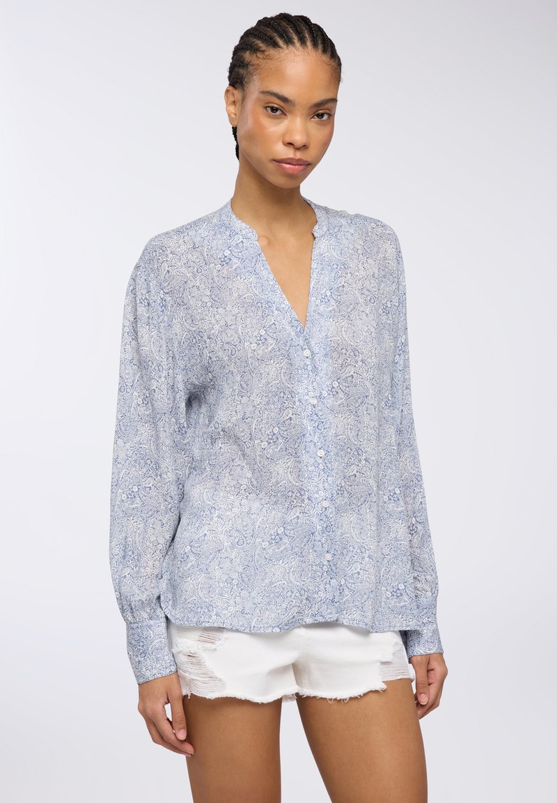 Camicia blu con motivo paisley, maniche lunghe, scollatura a V e chiusura con bottoni; tessuto leggero; indossata con pantaloni corti bianchi strappati.